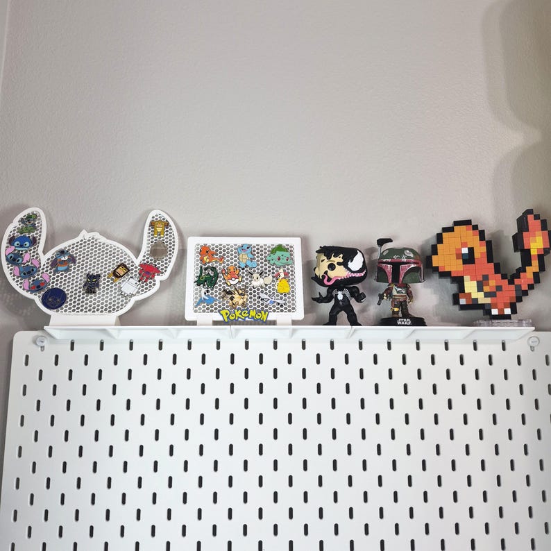Puede incluir: Un expositor de pines blanco con forma de Stitch, un tablero rectangular blanco con la palabra Pokemon, una figura negra de Venom, una figura de Boba Fett y una figura pixelada naranja de Charmander en un estante sobre un panel perforado blanco.