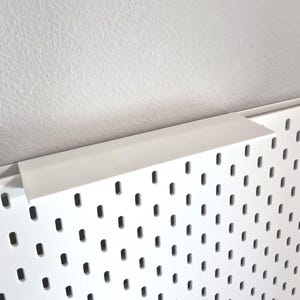 Puede incluir: Un panel perforado blanco con un patr&oacute;n repetitivo de ranuras verticales rectangulares redondeadas. En el borde superior hay un estante blanco s&oacute;lido con una superficie plana y un borde perpendicular que sobresale del tablero.