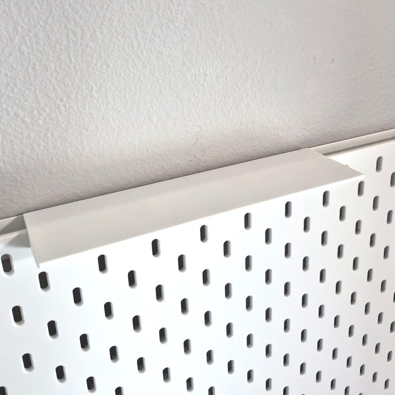 Puede incluir: Un panel perforado blanco con un patr&oacute;n repetitivo de ranuras verticales rectangulares redondeadas. En el borde superior hay un estante blanco s&oacute;lido con una superficie plana y un borde perpendicular que sobresale del tablero.