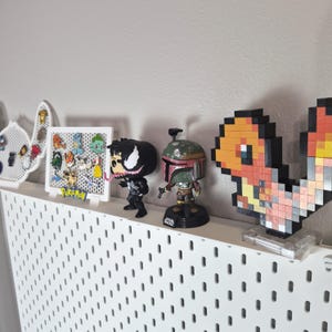 Puede incluir: Un estante de pared perforado blanco con una colecci&oacute;n de figuras y accesorios de cultura pop. Los art&iacute;culos incluyen un tablero expositor blanco con varios pines de personajes, una figura negra de Venom, una figura de Boba Fett y un Charmander pixelado.