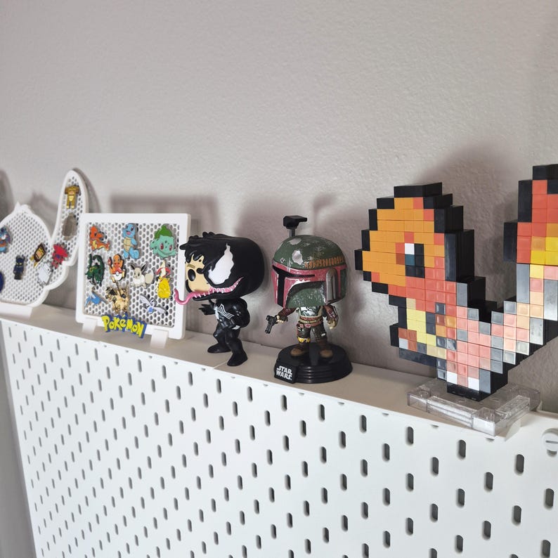 Puede incluir: Un estante de pared perforado blanco con una colecci&oacute;n de figuras y accesorios de cultura pop. Los art&iacute;culos incluyen un tablero expositor blanco con varios pines de personajes, una figura negra de Venom, una figura de Boba Fett y un Charmander pixelado.