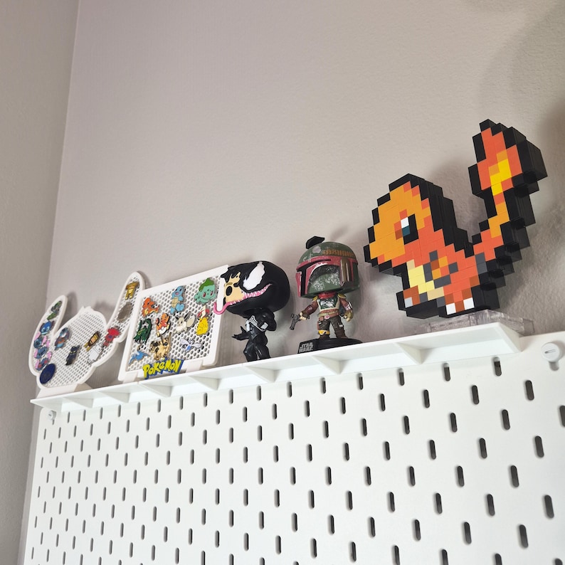 Puede incluir: Un estante de tablero perforado blanco muestra una colecci&oacute;n de art&iacute;culos: un tablero de pines blanco con forma de Stitch, un tablero de pines rectangular de Pokemon, una figura negra de Venom, una figura de Boba Fett y una figura pixelada de Charmander.