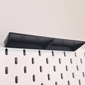 Puede incluir: Un estante rectangular negro dise&ntilde;ado para un tablero perforado blanco. El estante cuenta con una superficie horizontal plana con un panel trasero vertical y un divisor central, proporcionando dos secciones de almacenamiento distintas.