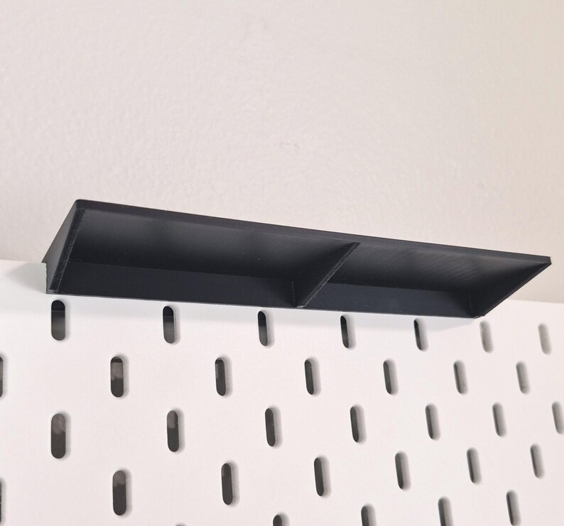 Puede incluir: Un estante rectangular negro dise&ntilde;ado para un tablero perforado blanco. El estante cuenta con una superficie horizontal plana con un panel trasero vertical y un divisor central, proporcionando dos secciones de almacenamiento distintas.