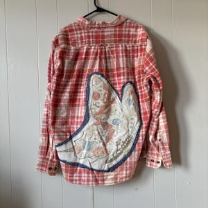 May include: A long-sleeve button-down shirt in a red and white plaid pattern with a bleached effect. The back features a large, asymmetrical fabric appliqué shaped like a butterfly wing, decorated with a floral print and a dark blue border.