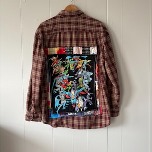 May include: A long-sleeved, button-down shirt in a maroon and tan plaid pattern. The back features a large, black patch with a colorful comic book illustration of various superheroes. Patches of fabric in different colors and patterns are sewn around the comic book patch.