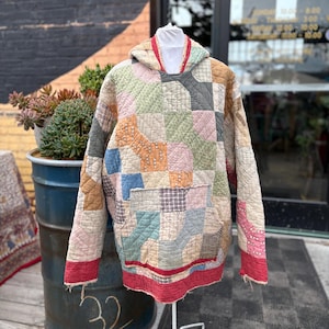 May include: A patchwork hoodie in a variety of colors, including beige, pink, blue, and green. The hoodie has a front pocket and a red trim along the bottom and hood. The garment is made of quilted fabric.