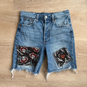 May include: Light blue denim shorts with distressed edges and two decorative patches. One patch features a floral design with skulls, and the other displays the text "Grateful Dead" with skeleton figures. The shorts have a button closure and classic five-pocket styling.