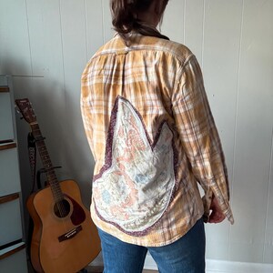 May include: A long-sleeved, button-down shirt in a plaid pattern of yellow, orange, and white. The back features a large, decorative patch with floral and paisley designs, bordered by a dark purple trim. The shirt is worn with blue jeans.