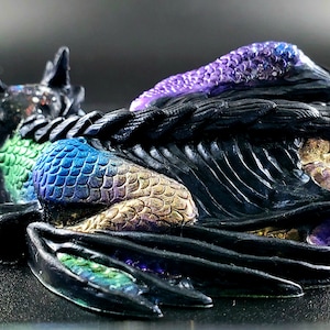 Peut inclure: Une figurine de dragon noire, verte, dorée et violette avec des ailes déployées. Le dragon est enroulé sur lui-même dans une position de sommeil.