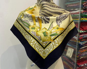 May include: A cream-colored silk scarf with a black border and a floral pattern. The pattern includes yellow flowers, green leaves, and a gray bird. The scarf is displayed on a mannequin.