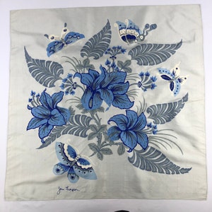 May include: A square, pale grey scarf with a floral and butterfly design. The design features large blue flowers, grey leaves, and blue butterflies. The scarf has the text "Jim Thompson" in the bottom left corner.