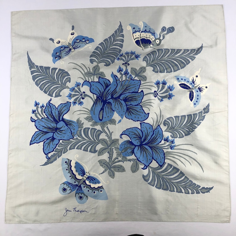 May include: A square, pale grey scarf with a floral and butterfly design. The design features large blue flowers, grey leaves, and blue butterflies. The scarf has the text "Jim Thompson" in the bottom left corner.