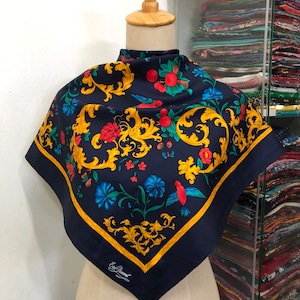 May include: A navy blue silk scarf with a gold, red, and green floral pattern. The scarf features a repeating design of birds, flowers, and leaves. The scarf is trimmed with a gold border and has the text "Eric Bompard" in the lower right corner.