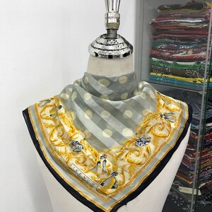 May include: A square silk scarf with a gray and white striped pattern and large white polka dots. The scarf has a gold floral border with the word "GIVENCHY" printed on it. The scarf is displayed on a mannequin.