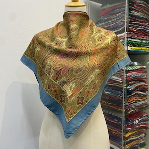 May include: A square silk scarf featuring an intricate paisley pattern in shades of olive green, gold, and pink, bordered by a solid dusty blue edge. The word LIBERTY is printed in a small rectangular box on the lower section of the fabric.
