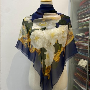 Può includere: Una sciarpa quadrata di seta con bordo blu navy e motivo floreale. La sciarpa presenta grandi fiori bianchi con centri gialli e foglie verdi. Cornici dorate ornate circondano la composizione floreale. La sciarpa è drappeggiata su un manichino.