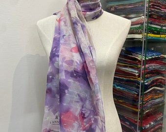 Könnte beinhalten: Ein weißer Seidenschal mit einem Aquarell-Blumenmuster in Violett-, Rosa- und Grautönen. Der Schal trägt den Schriftzug "LANVIN COLLECTION" in Schwarz.