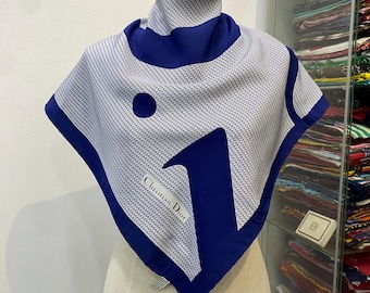 Könnte beinhalten: Ein quadratisches Seidentuch mit blau-weißem Muster, präsentiert auf einer Schaufensterpuppe. Das Tuch zeigt einen großen blauen Buchstaben "V" und ein kreisförmiges Designelement. Die Kanten sind blau eingefasst, und der Markenname "Christian Dior" ist sichtbar.
