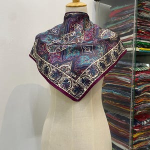 May include: A square scarf featuring a complex paisley and floral pattern in shades of purple, teal, and cream. The scarf has a dark purple border and the word LIBERTY is printed in a small white box near the bottom edge of the fabric design.