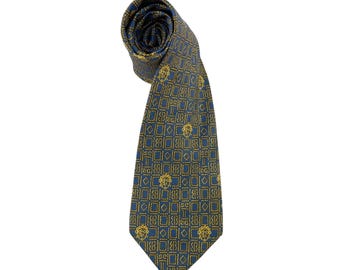 May include: A rolled-up blue tie with a repeating gold geometric pattern and a central face design. The tie is made of a shiny material and is set against a white background.