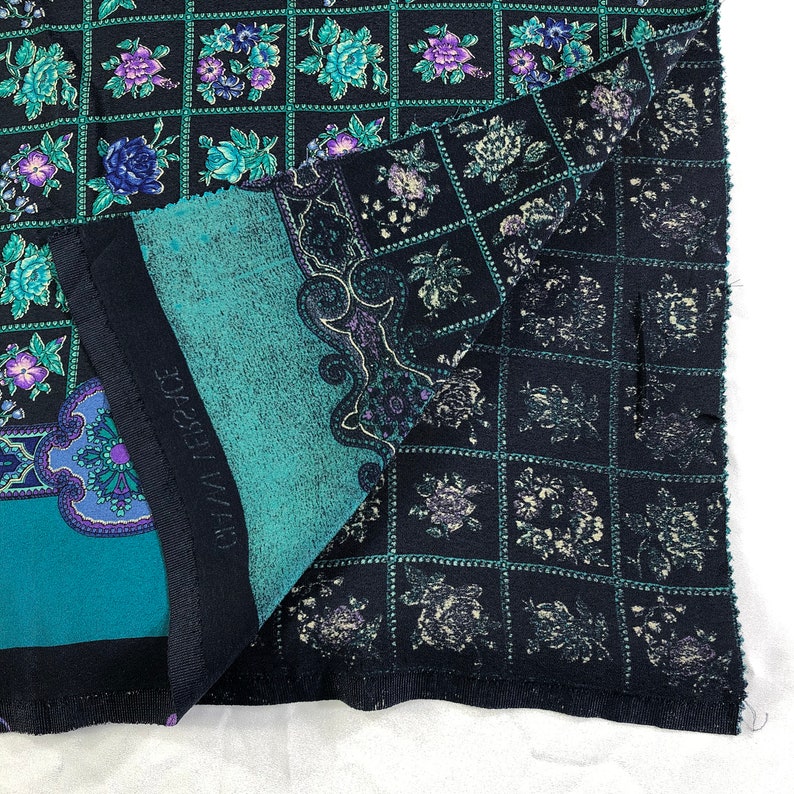 May include: A black and teal fabric with a repeating pattern of floral designs. The fabric has a grid pattern with a floral design in each square. The fabric is folded in half and the text "MADE IN FRANCE" is visible.