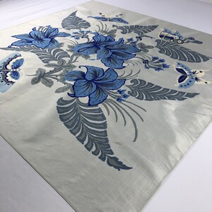 May include: A square, pale grey scarf with a floral and butterfly design. The scarf features large blue flowers and grey leaves and stems. Butterflies in shades of blue and white are also present, creating a nature-inspired pattern.