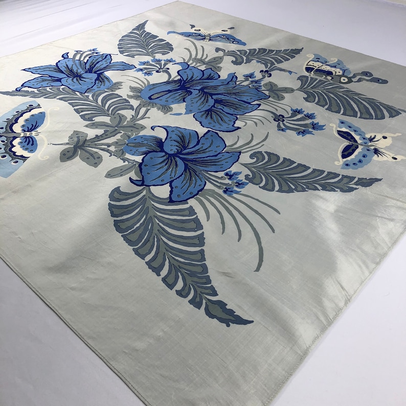May include: A square, pale grey scarf with a floral and butterfly design. The scarf features large blue flowers and grey leaves and stems. Butterflies in shades of blue and white are also present, creating a nature-inspired pattern.