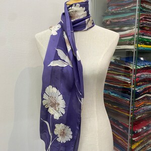 May include: A purple silk scarf with a white floral print. The scarf is tied around a white mannequin's neck.