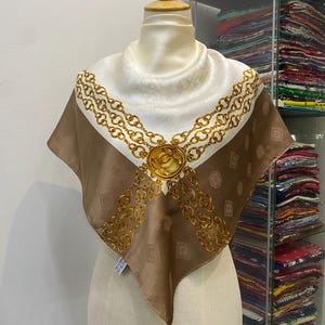 Può includere: Un foulard quadrato di seta bianco e marrone, ornato da un motivo a catena dorata e da un medaglione centrale. Il foulard è esposto su un manichino, che mette in risalto la sua elegante drappeggio e il suo motivo intricato. Il design della sciarpa presenta un motivo ripetuto di piccoli quadrati.