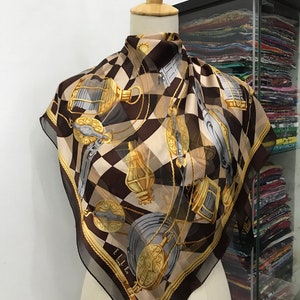 May include: A brown and beige silk scarf with a checkered pattern and gold accents. The scarf features a repeating design of gold chains, pulleys, and lanterns. The scarf is made by Elle Italia.