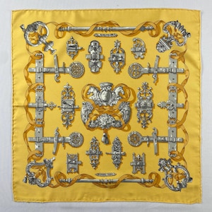 May include: A square yellow silk scarf featuring a repeating pattern of silver-toned metal door hardware, latches, and ornate knockers. The design includes a central crest with two horses and a lion head, all connected by flowing golden ribbons.