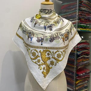 May include: A square, white silk scarf with a detailed illustration of horse-drawn carriages and figures in blue and brown. The scarf has a gold border and a central design with a golden lion and floral elements. The scarf is displayed on a mannequin.