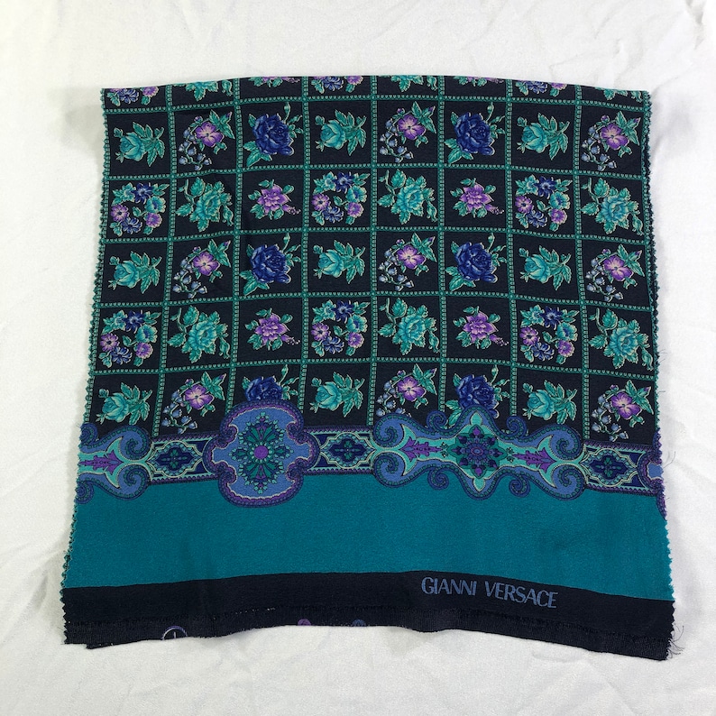 May include: A teal and black fabric with a floral pattern and a border. The fabric has the text "GIANNI VERSACE" printed on the bottom.