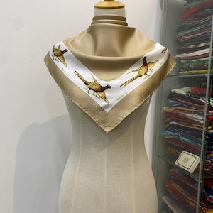 May include: A tan silk scarf with a white border featuring pheasant illustrations. The scarf is draped on a mannequin. The pheasant illustrations are in shades of brown, green, and red. The word "GUCCI" is printed on the scarf.