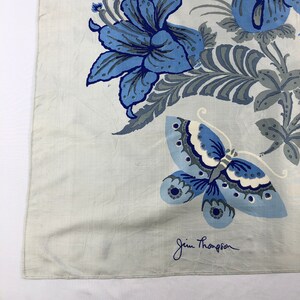 May include: A pale blue textile with a floral and butterfly pattern in shades of blue and grey. The design features large flowers and a butterfly with detailed wing patterns. The Jim Thompson signature is in the bottom right corner.