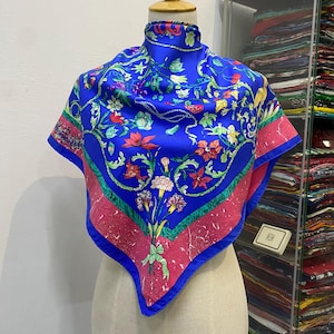 Könnte beinhalten: Ein leuchtend blauer Seidenschal mit floralem Design, auf einer Schaufensterpuppe präsentiert. Der Schal hat ein zentrales Muster aus bunten Blumen und Blättern, umrahmt von einem rosa Rand und blauen Kanten. Der Schal ist quadratisch.