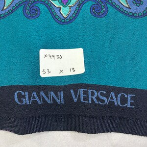 May include: A teal blue fabric with a paisley pattern and a black band with the text "GIANNI VERSACE" in blue. A white tag with the text "x4930 53 x 13" is attached to the fabric.