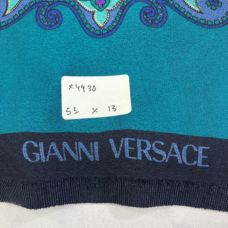 May include: A teal blue fabric with a paisley pattern and a black band with the text "GIANNI VERSACE" in blue. A white tag with the text "x4930 53 x 13" is attached to the fabric.