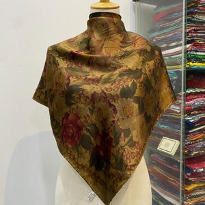 Può includere: Foulard quadrato in seta con motivo floreale nei toni del marrone, oro e bordeaux. Il foulard è drappeggiato su un manichino. I bordi presentano un motivo ripetuto. Misura circa 80 cm x 80 cm.