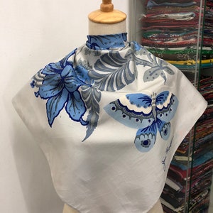 May include: A square, off-white silk scarf with a butterfly and floral design in shades of blue and grey. The scarf is displayed on a mannequin. The artist's signature is visible in the lower right corner.