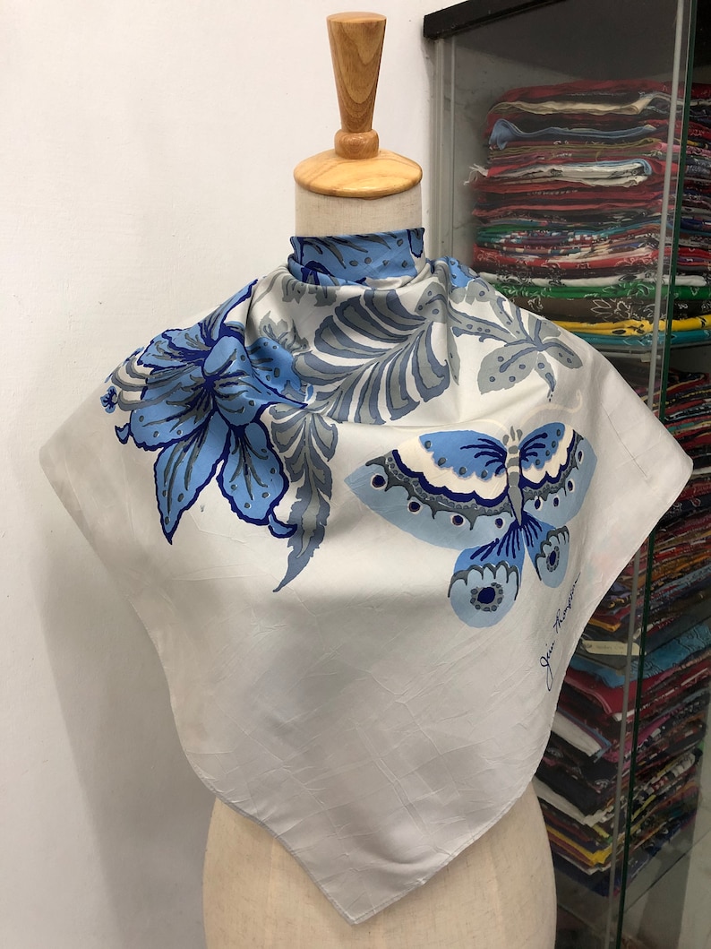 May include: A square, off-white silk scarf with a butterfly and floral design in shades of blue and grey. The scarf is displayed on a mannequin. The artist's signature is visible in the lower right corner.