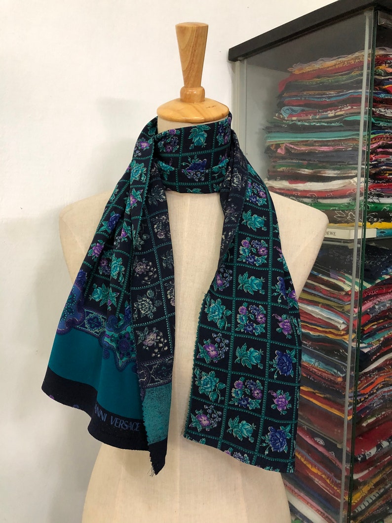 May include: A teal and black silk scarf with a floral pattern and the text "ANN VERSACE" printed on the bottom edge.