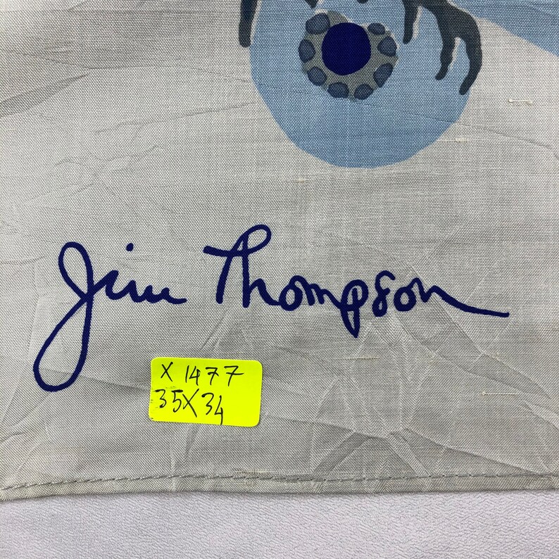 May include: A light grey Jim Thompson scarf featuring a blue floral pattern and the brand's signature in dark blue. The scarf has a yellow tag with the measurements 89 x 86 cm.