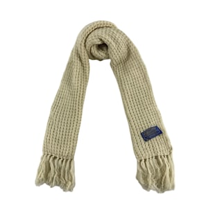 Può includere: Una sciarpa di lana color crema con frange alle estremità. La sciarpa ha una trama tessuta e un'etichetta blu con testo bianco che dice "Woolrich".