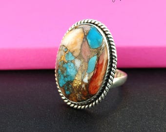 May include: A silver ring with a large oval-shaped gemstone. The gemstone is a mix of turquoise, coral, and other colorful stones. The ring has a rope-like design around the edge of the setting.