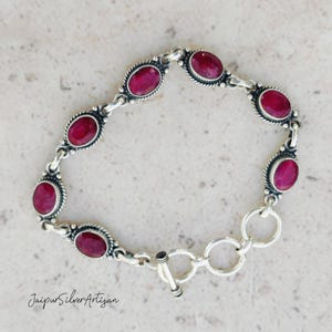 May include: A silver bracelet with a toggle clasp features oval-shaped red gemstones set in silver bezels. The bracelet is approximately 7 inches long.