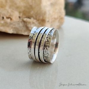 May include: A silver-colored spinner ring with a textured design. The ring features multiple bands, some with etched patterns. The ring is displayed on a light-colored surface, with a blurred background.