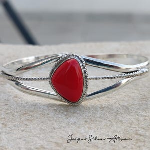 May include: A silver cuff bracelet featuring a vibrant red, triangular gemstone at its center. The bracelet has a twisted rope design along the edges and is set against a neutral background. The text "Jaipur Silver Artisan" is at the bottom.