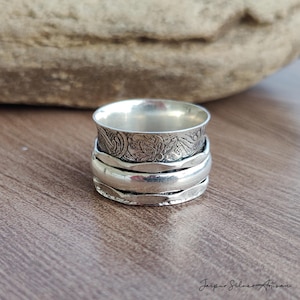 May include: A silver spinner ring with a floral design on the top band. The ring has multiple bands that spin freely. The ring is on a wooden surface. The text "Jaipur Silver Artisan" is in the bottom right corner.